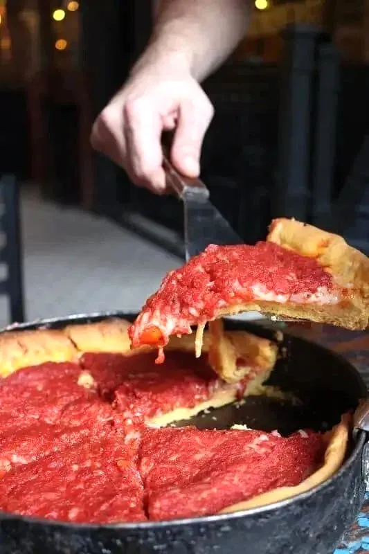 Hands stretching melted cheese from deep-dish pizza slice over cast iron pan