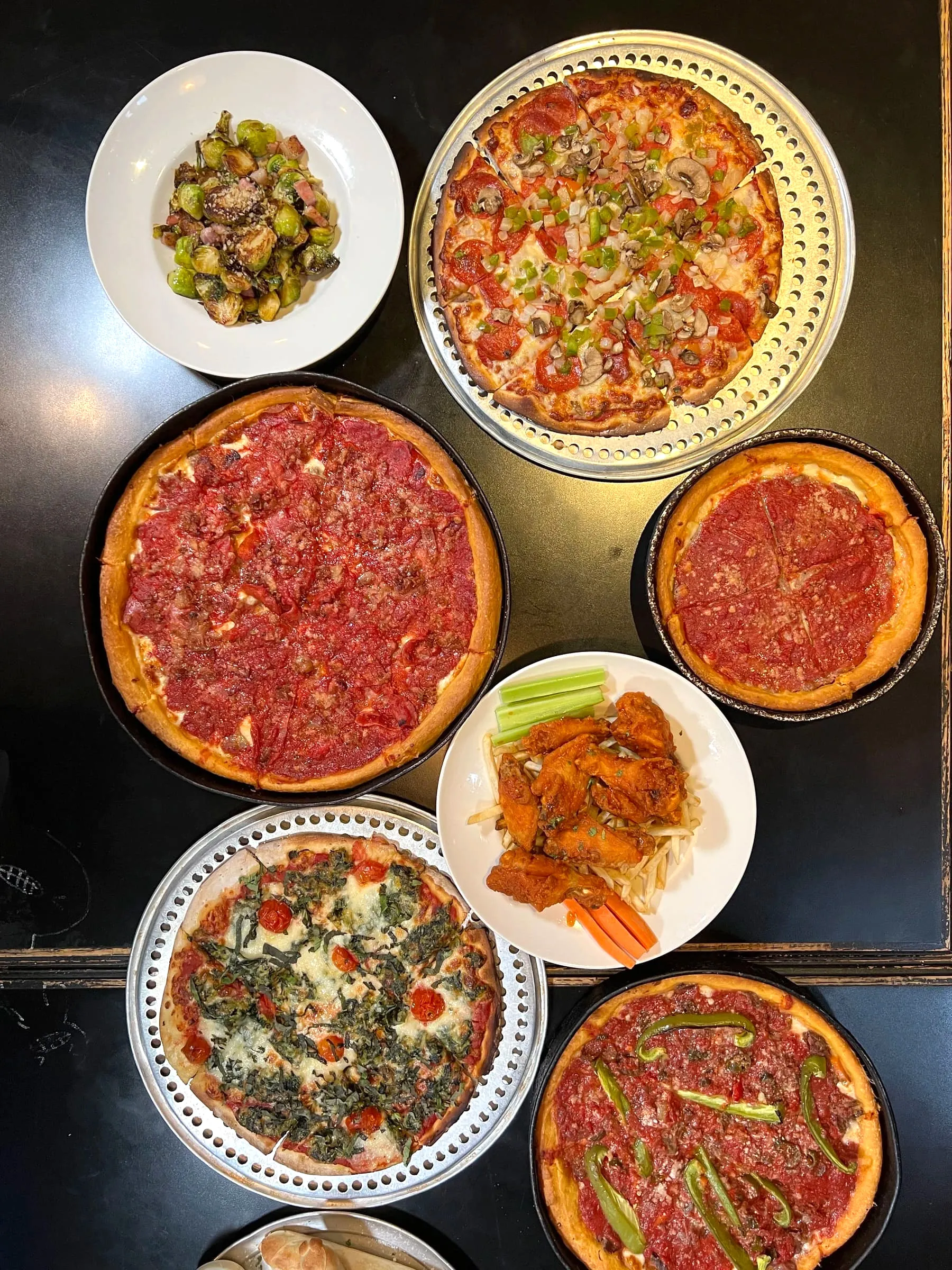 Overhead view of a catering spread with multiple deep dish and thin crust pizzas, wings, and salads