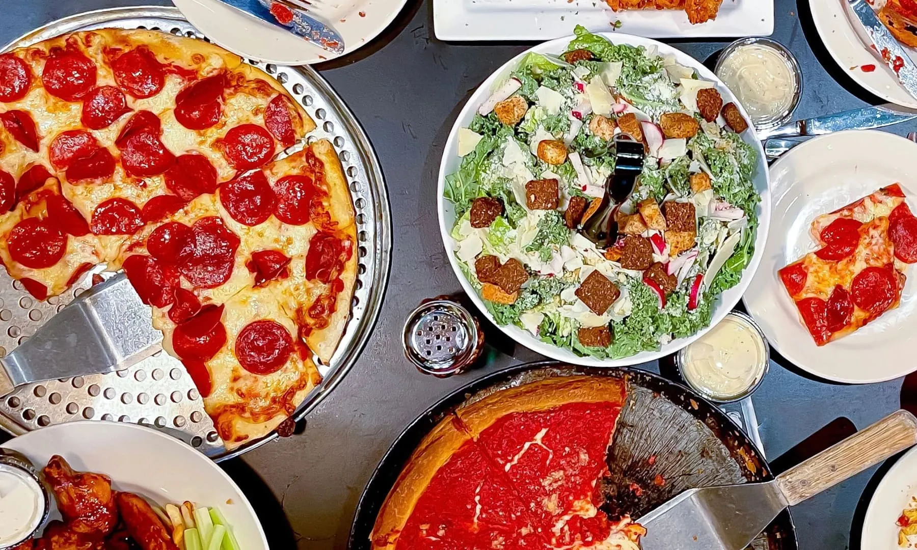 Overhead spread of pepperoni pizza, Caesar salad, chicken wings, and deep dish pizza for catering